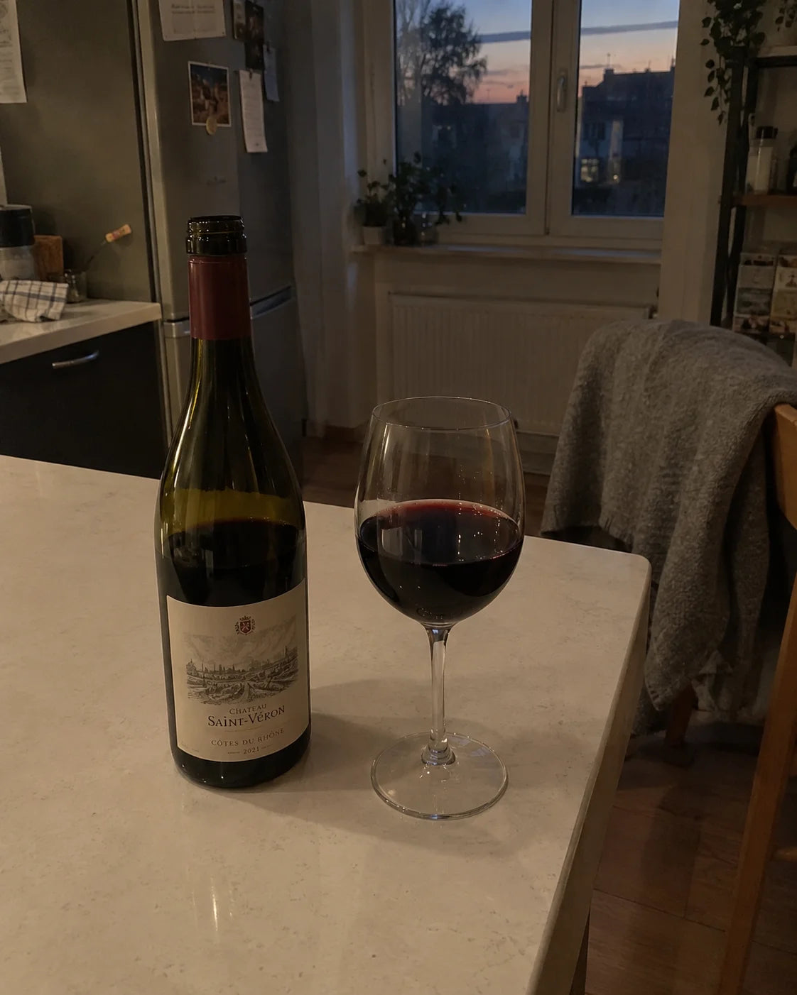 A bottle of wine and a poured glass on a kitchen counter at dusk, a cardigan draped over a chair