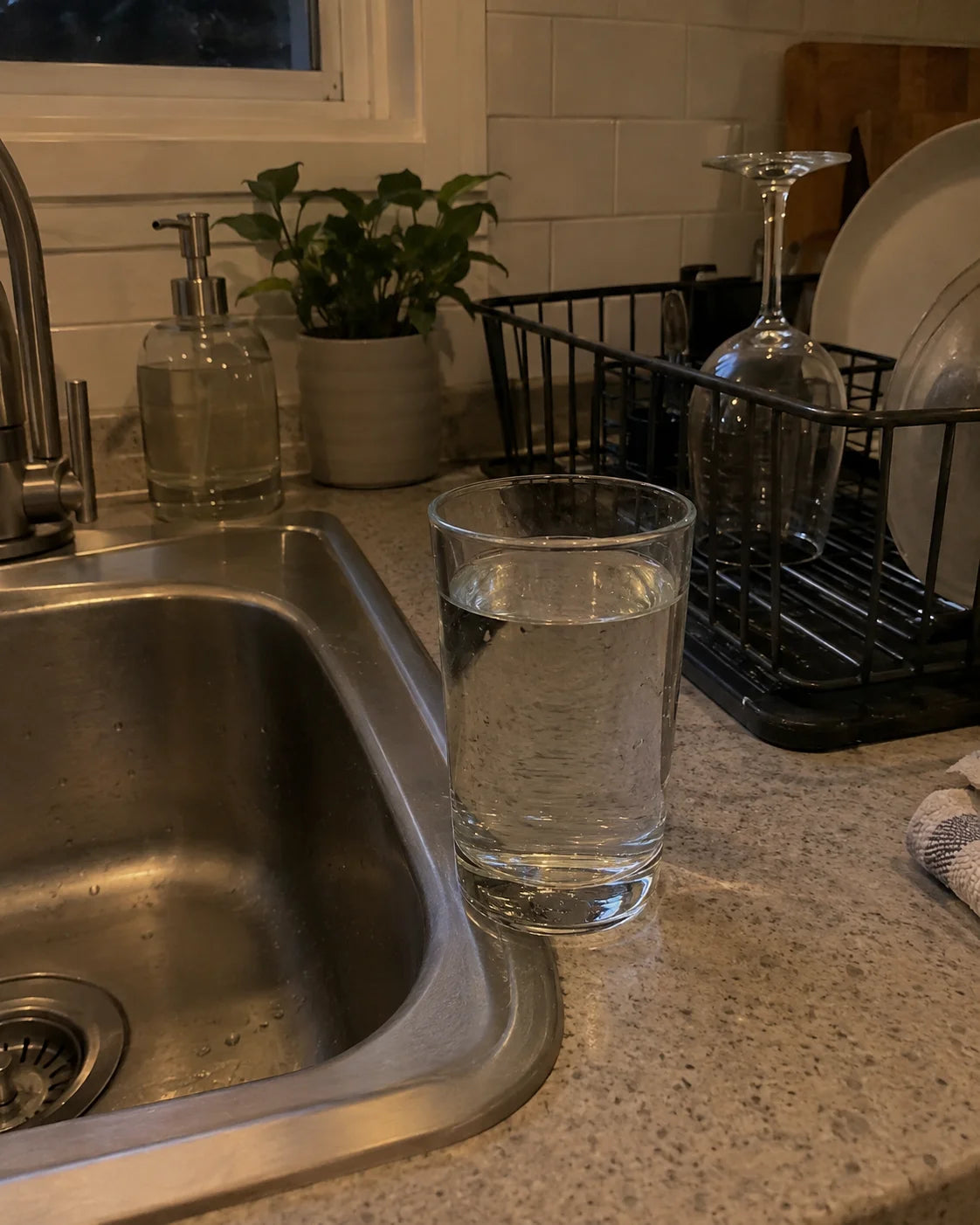 A glass of water by the kitchen sink in the evening, an empty wine glass turned upside down in the drying rack