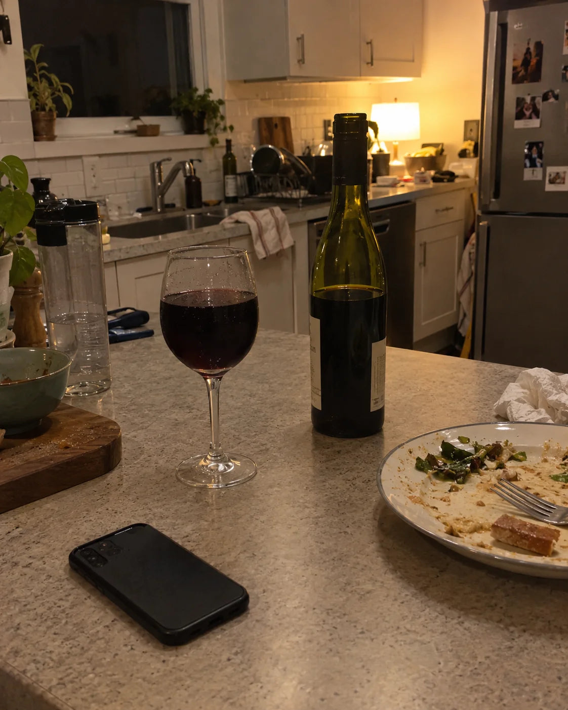 An almost empty wine bottle next to a full glass on a kitchen counter at night