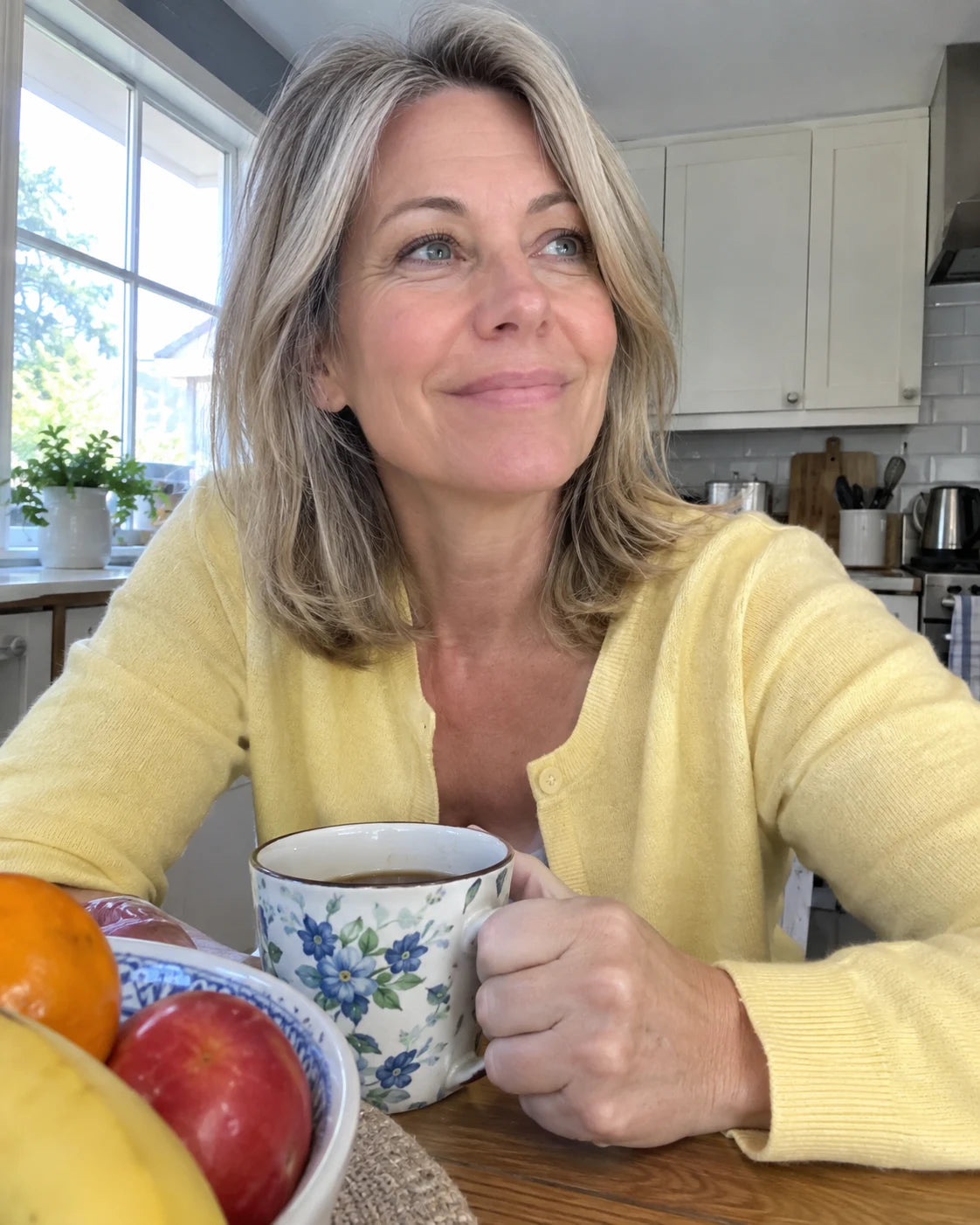Woman smiling at her kitchen table in the morning light holding a mug of tea