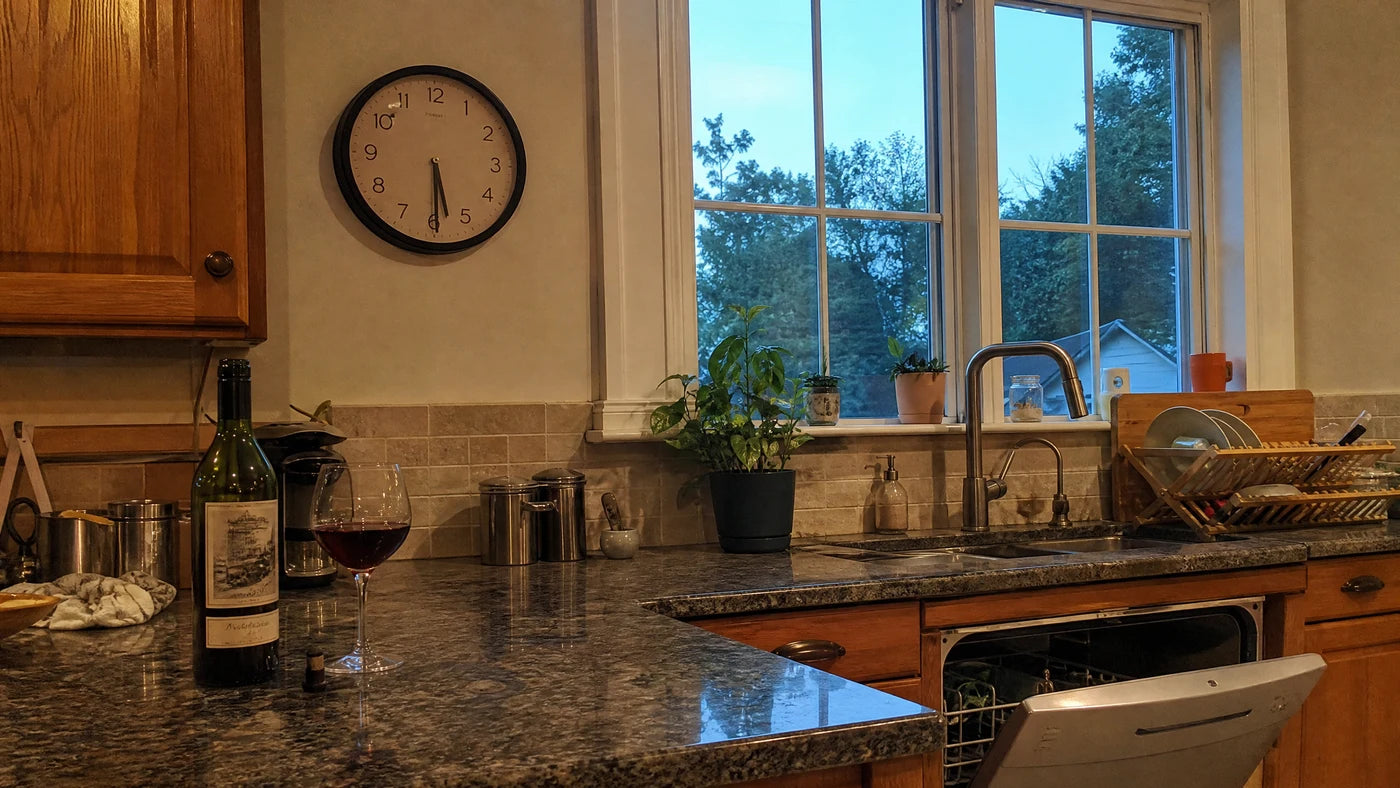 A kitchen counter in the early evening with a wall clock reading just before 6 o'clock and a freshly poured glass of wine
