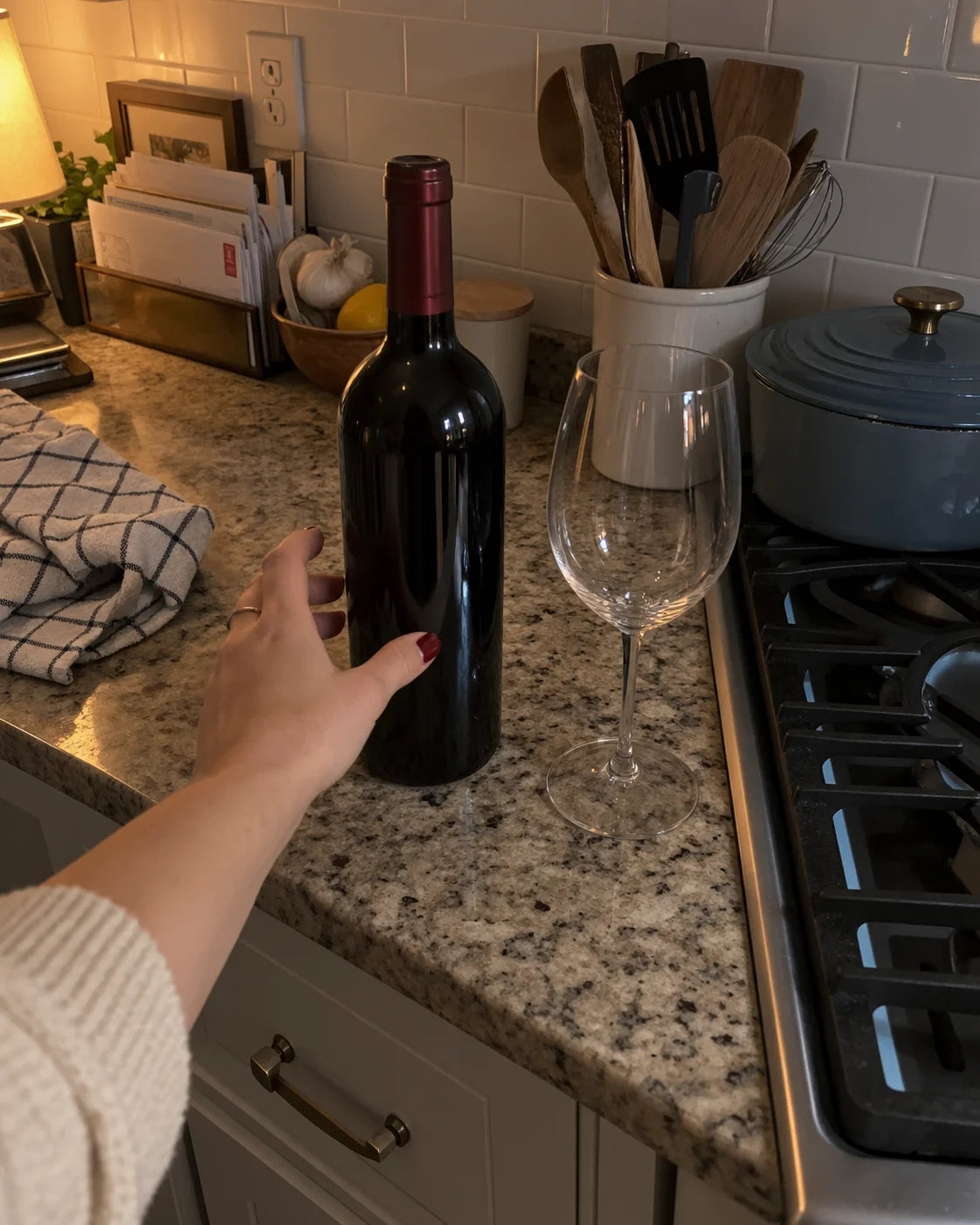 A hand reaching for a wine bottle on a kitchen counter next to an empty glass at dusk