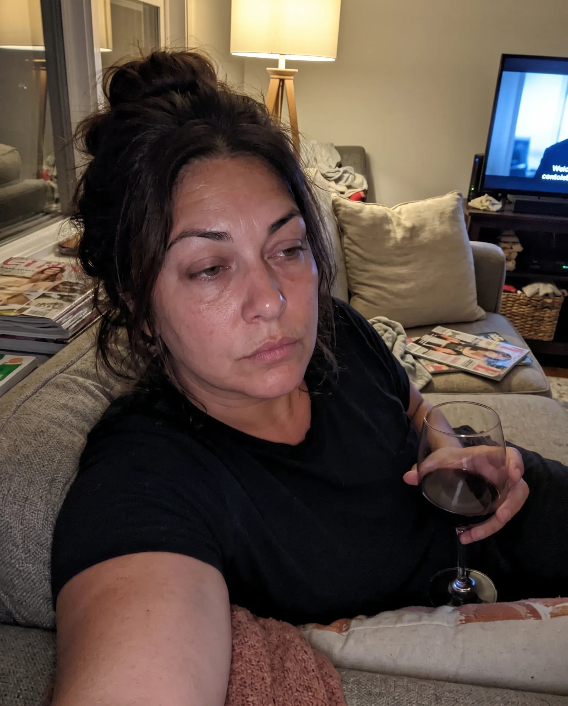 A woman on her couch at night holding a glass of wine with a flat, tired expression