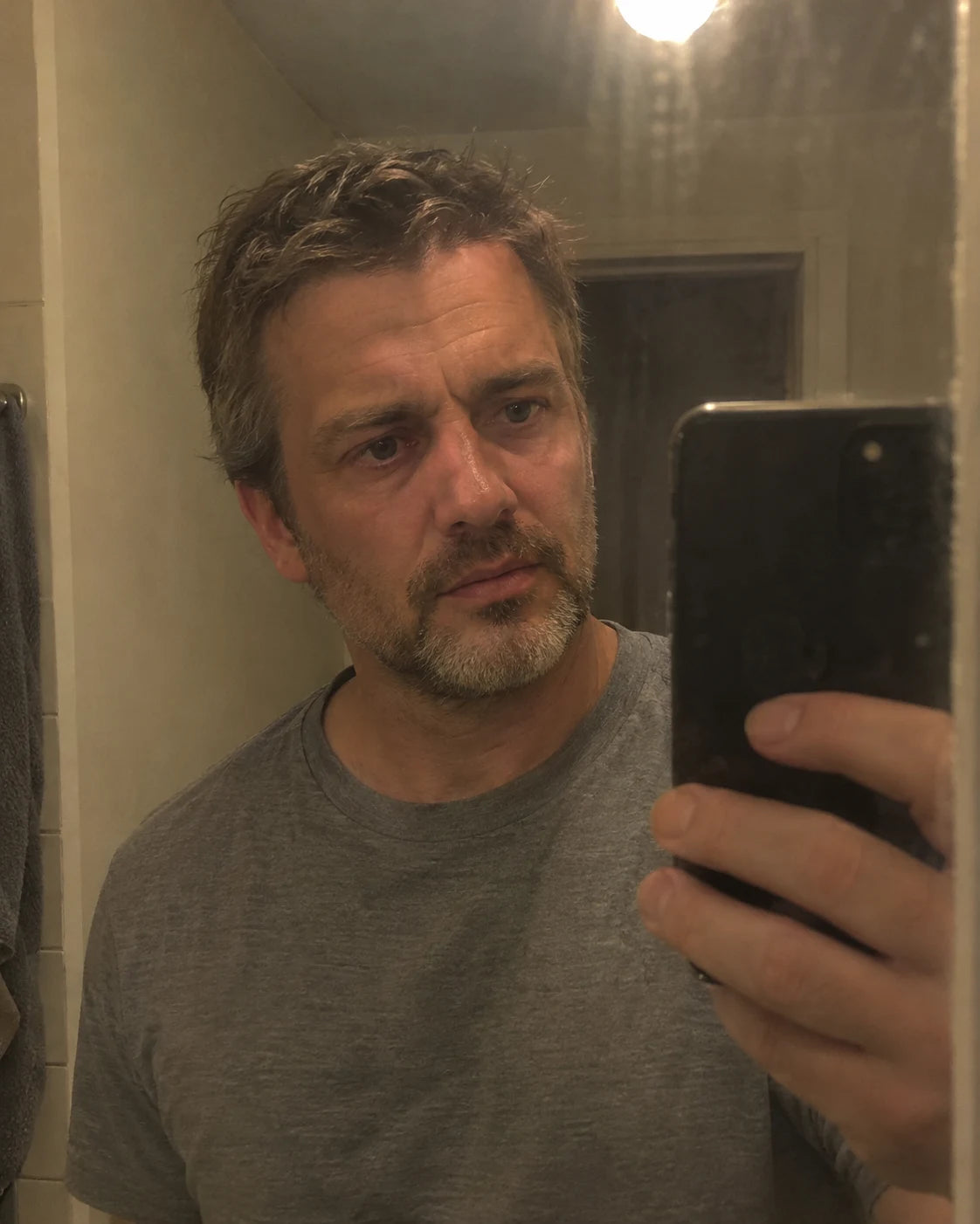 A man looking tiredly at his own reflection in a bathroom mirror at night