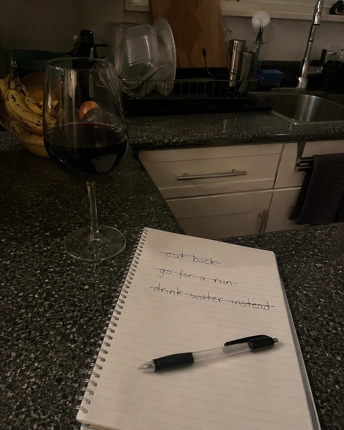A full glass of red wine next to an open notebook with a crossed-out to-do list on a kitchen counter at night