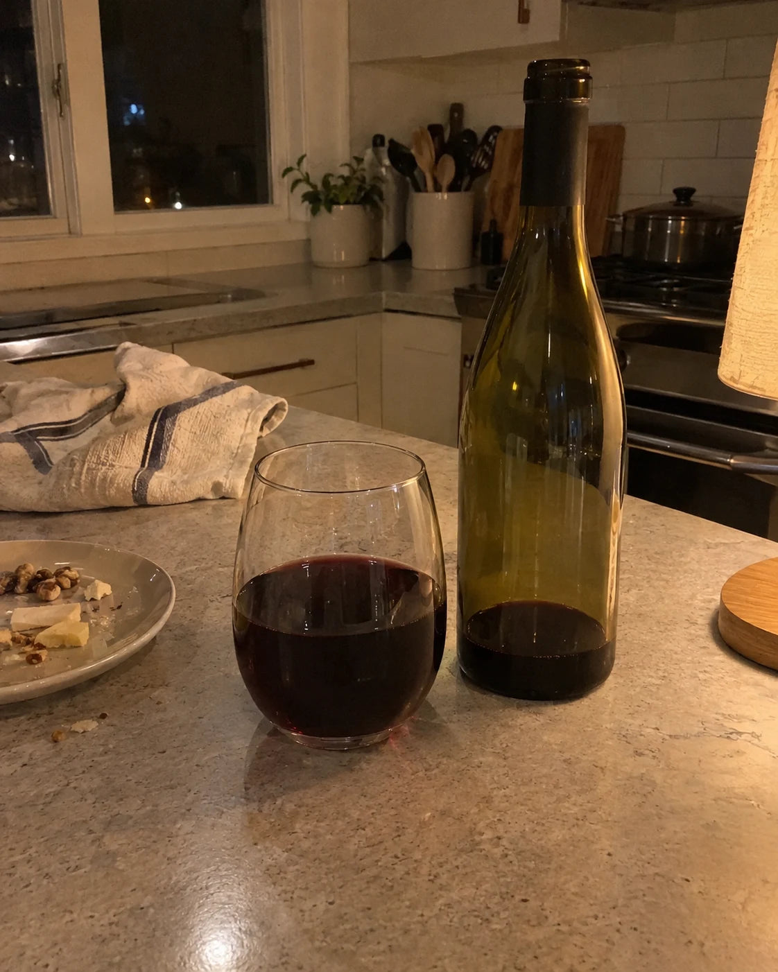 A glass of red wine and a nearly empty bottle on a kitchen counter at night