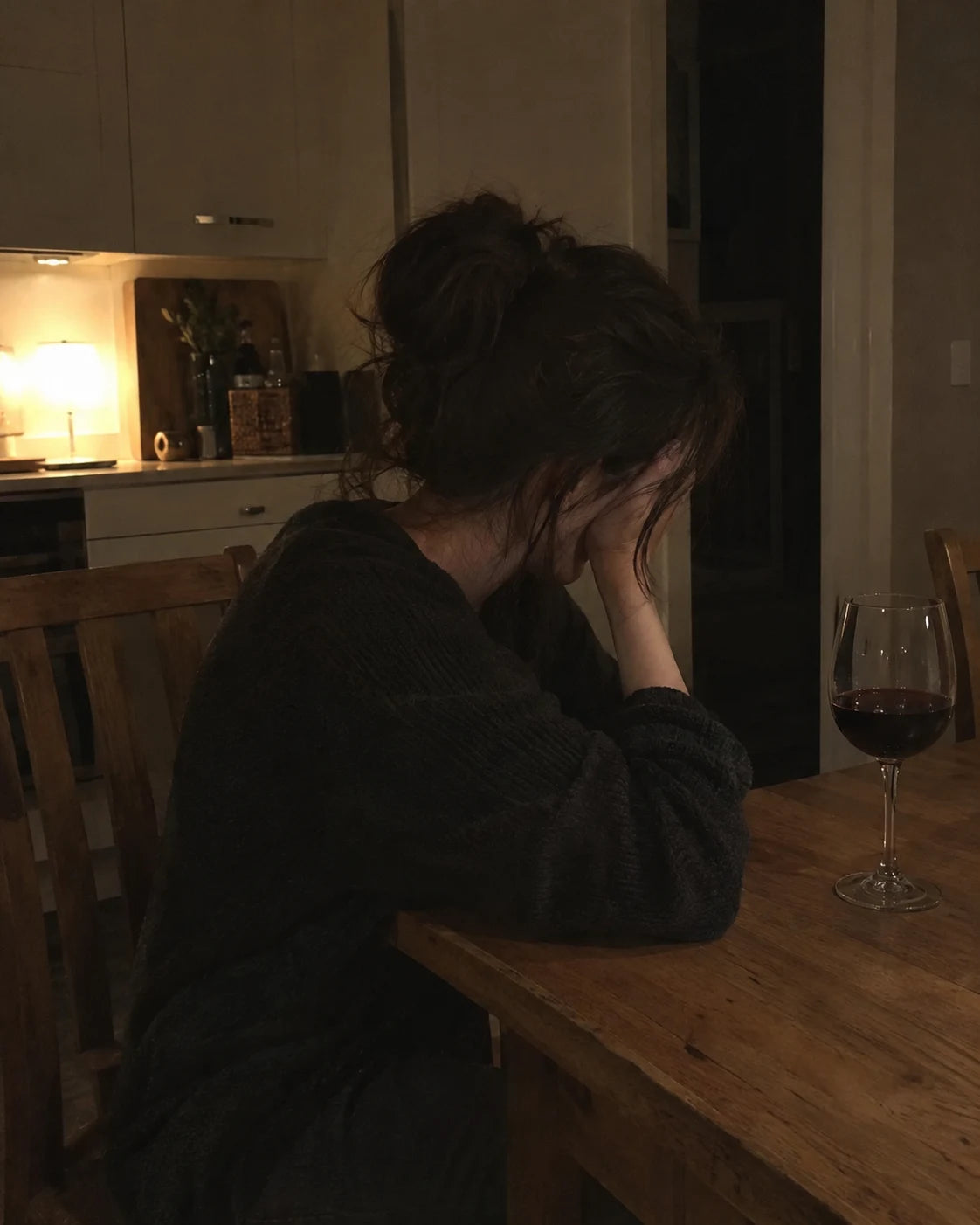 A woman slumped alone at a kitchen table late at night, head in hand, next to an untouched glass of wine