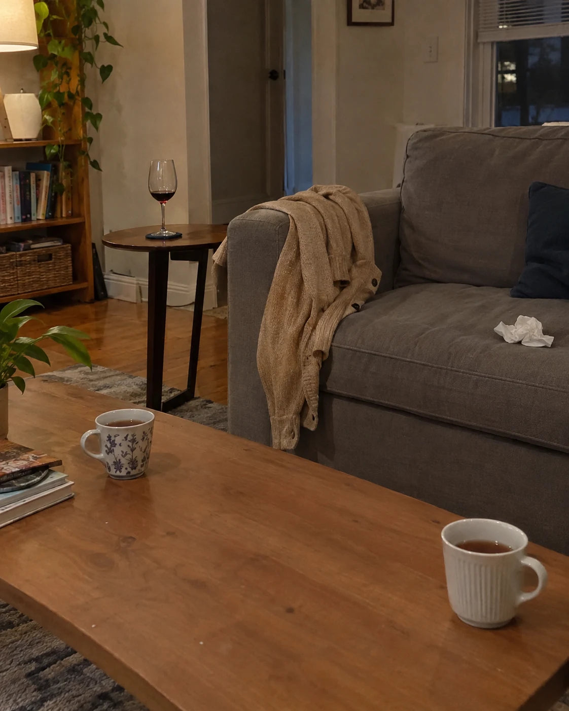 A glass of untouched red wine and two cooling mugs of tea sitting far apart in an empty living room, a cardigan draped over the couch, a crumpled tissue on the cushion