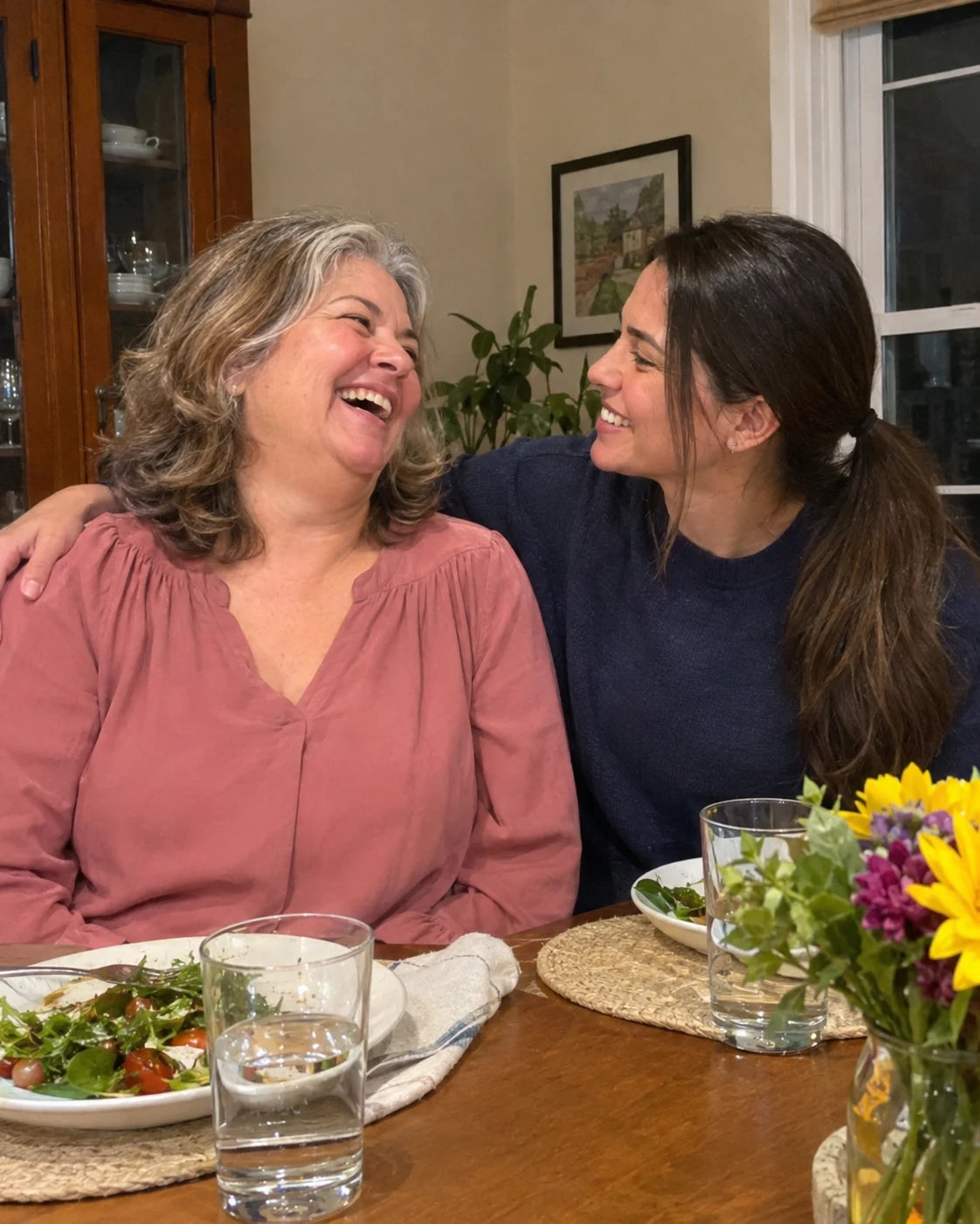 An older woman laughing warmly with her adult daughter's arm around her shoulders at a dinner table