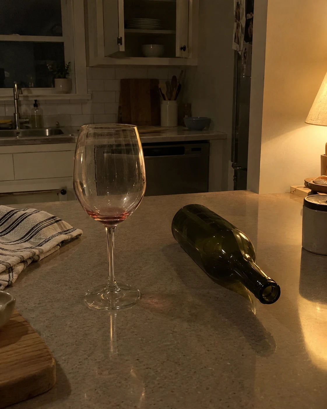 An empty wine glass next to an empty bottle on a dim kitchen counter at night