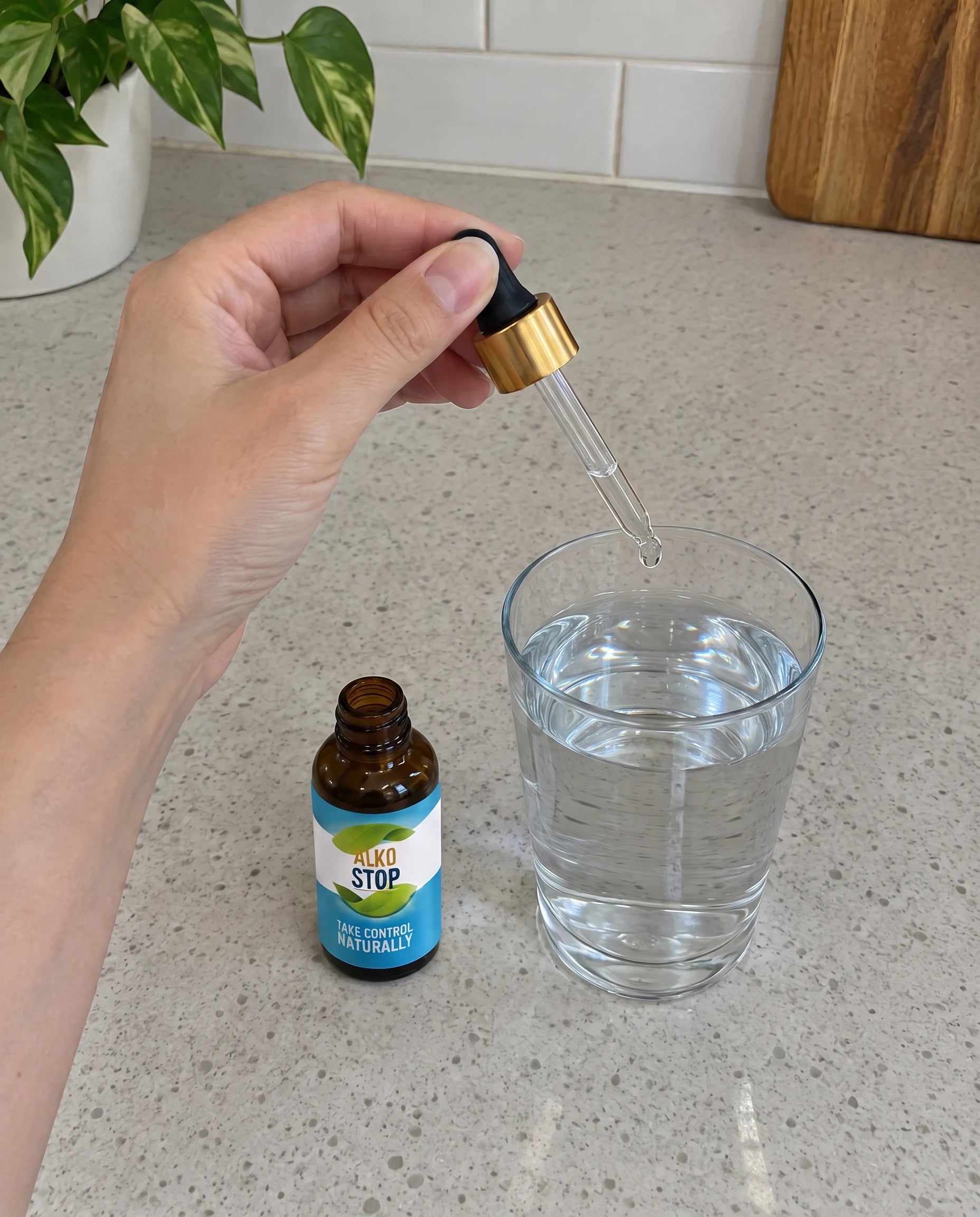 An AlkoStop bottle resting on the counter, one hand holding the dropper with its gold collar above a glass of water