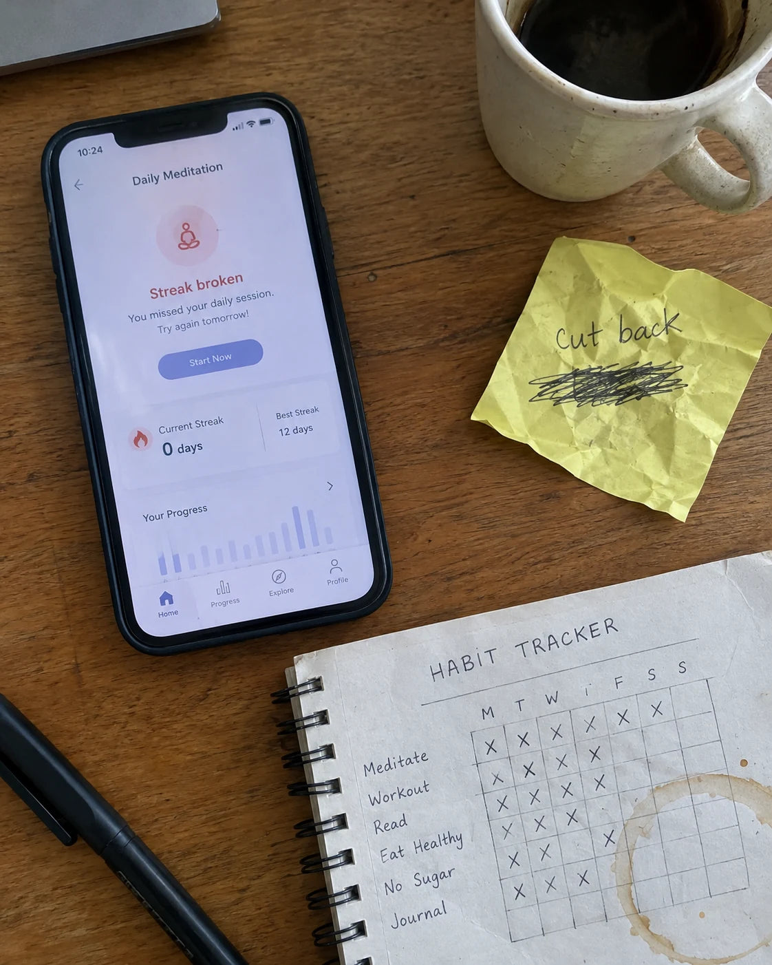 A phone showing a broken meditation streak next to a crossed-out habit tracker notebook