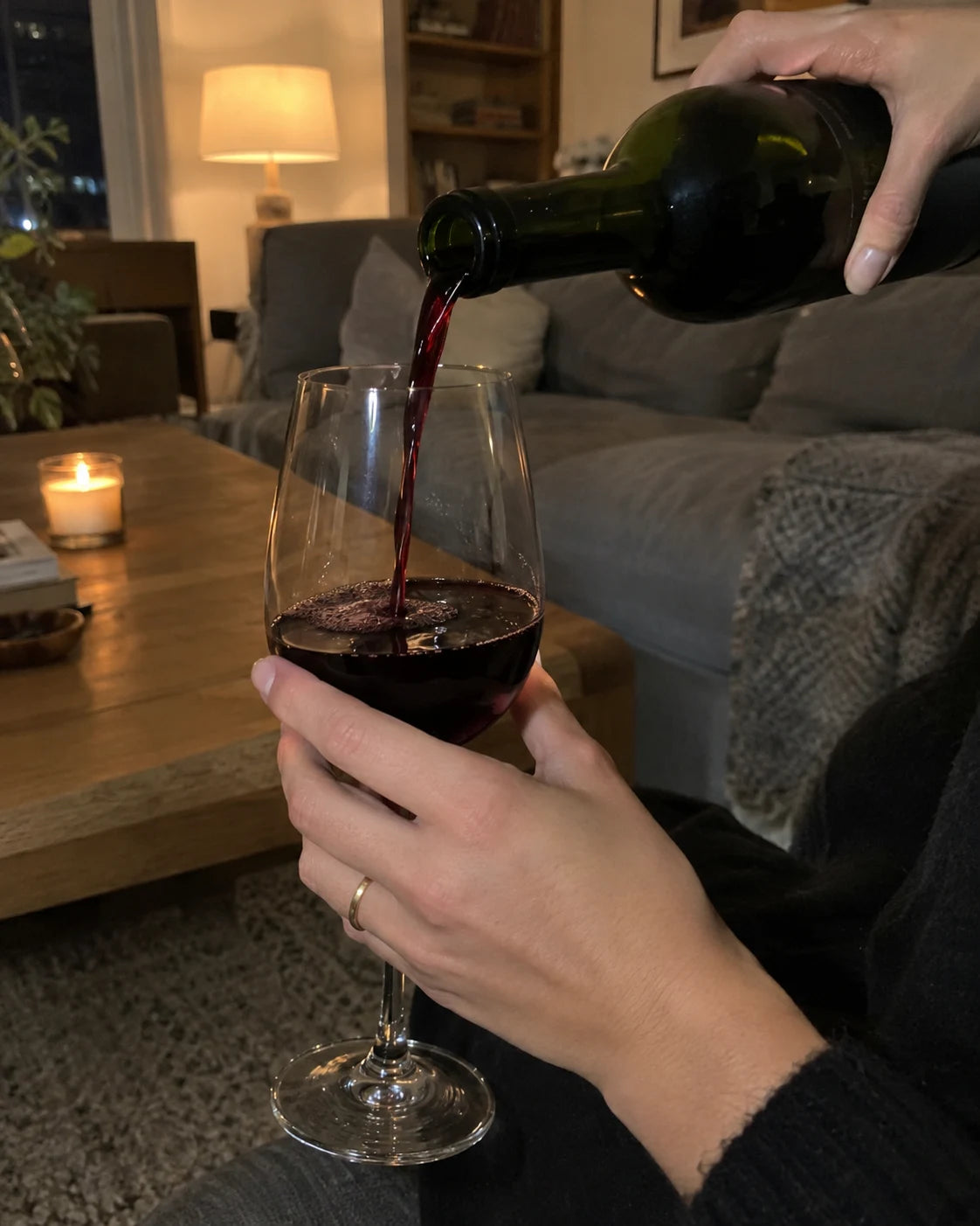 Hand pouring red wine into a glass in a cozy living room, evening